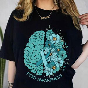 May include: Black t-shirt featuring a teal brain graphic on the left and a floral arrangement on the right. A teal ribbon and the words "PTSD Awareness" are also displayed. The design is centered on the shirt.