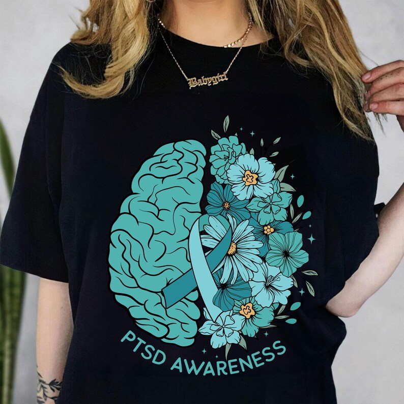 May include: Black t-shirt featuring a teal brain graphic on the left and a floral arrangement on the right. A teal ribbon and the words "PTSD Awareness" are also displayed. The design is centered on the shirt.