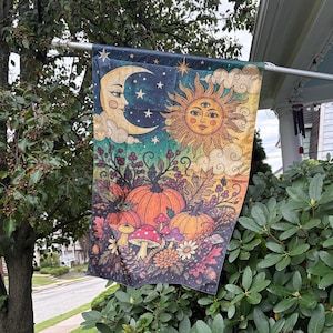 May include: A rectangular garden flag featuring a celestial design with a crescent moon and a sun with three eyes. Below, the illustration displays orange pumpkins, red and white spotted mushrooms, autumn leaves, and berries on a dark background.