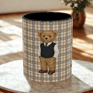 May include: A cylindrical container with a black interior and a plaid pattern in shades of beige, gray, and white. A cartoon bear wearing a vest, shirt, and pants is featured on the front. The container is sitting on a marble table.