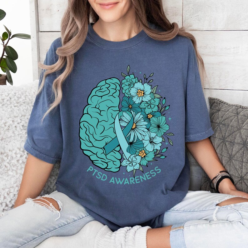 May include: A blue t-shirt featuring a teal brain graphic on the left side, with teal flowers and a teal ribbon on the right. The words "PTSD Awareness" are printed in teal below the graphics. The shirt is a solid blue color.