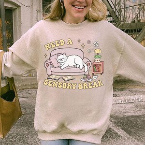 May include: Beige sweatshirt with a graphic of a white cat lounging on a pink couch. The text "NEED A SENSORY BREAK" is arched above and below the couch. The design includes books, a lamp, and a small table.