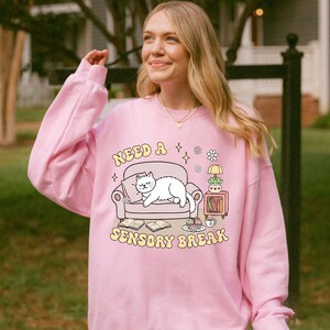 May include: A light pink sweatshirt with the text "Need a Sensory Break" and a graphic of a cat on a couch. The graphic also includes books, a side table, and a cup. The sweatshirt is a soft, comfortable material.