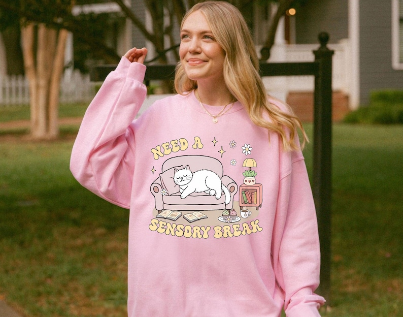 May include: A light pink sweatshirt with the text "Need a Sensory Break" and a graphic of a cat on a couch. The graphic also includes books, a side table, and a cup. The sweatshirt is a soft, comfortable material.