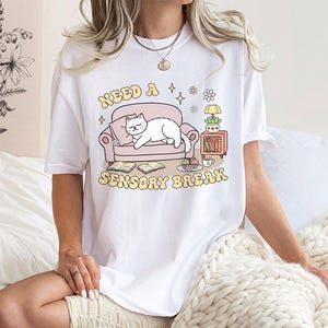 May include: White t-shirt with a graphic that reads "Need a Sensory Break." The design features a cartoon cat lounging on a pink couch, with a side table and books. The text and graphics are in soft pastel colors.