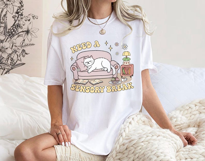 May include: White t-shirt with a graphic that reads "Need a Sensory Break." The design features a cartoon cat lounging on a pink couch, with a side table and books. The text and graphics are in soft pastel colors.