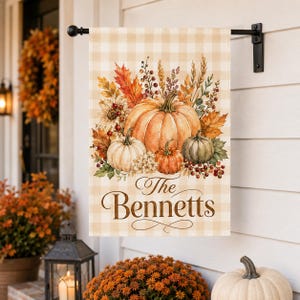 May include: A rectangular garden flag featuring a light beige and white buffalo check pattern. The design displays a cluster of orange, white, and green pumpkins, autumn leaves, wheat stalks, and red berries. The text The Bennetts is printed.