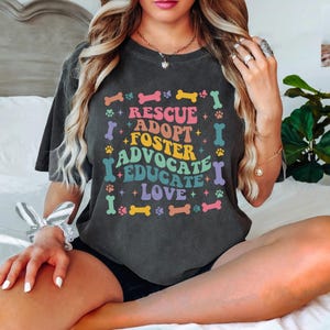 May include: Dark grey t-shirt featuring the words "RESCUE, ADOPT, FOSTER, ADVOCATE, EDUCATE, LOVE" in a colourful, retro font. The design incorporates paw prints and bone shapes. The shirt has a relaxed fit.