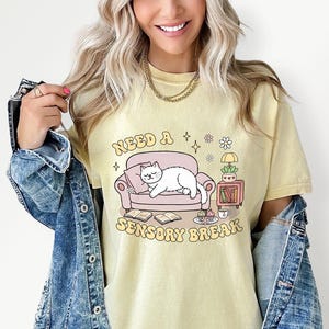 May include: A light yellow t-shirt with the text "Need a Sensory Break" and a graphic of a white cat lounging on a pink couch. The graphic also includes books, a lamp, and a small table with a plant and a cup.
