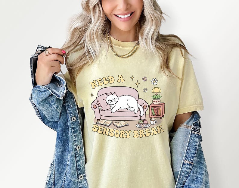 May include: A light yellow t-shirt with the text "Need a Sensory Break" and a graphic of a white cat lounging on a pink couch. The graphic also includes books, a lamp, and a small table with a plant and a cup.