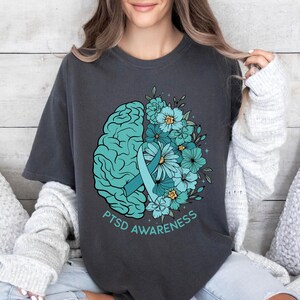 May include: Dark gray t-shirt featuring a teal brain graphic with a teal ribbon and teal flowers. The words "PTSD Awareness" are printed in teal below the graphic. The shirt is a comfortable, casual style.