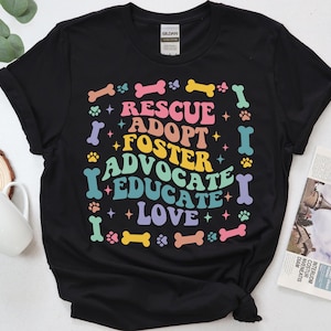 May include: Black t-shirt featuring the words "RESCUE, ADOPT, FOSTER, ADVOCATE, EDUCATE, LOVE" in a vibrant, retro style. The design incorporates paw prints, bones, and stars. The shirt is made of a soft fabric.