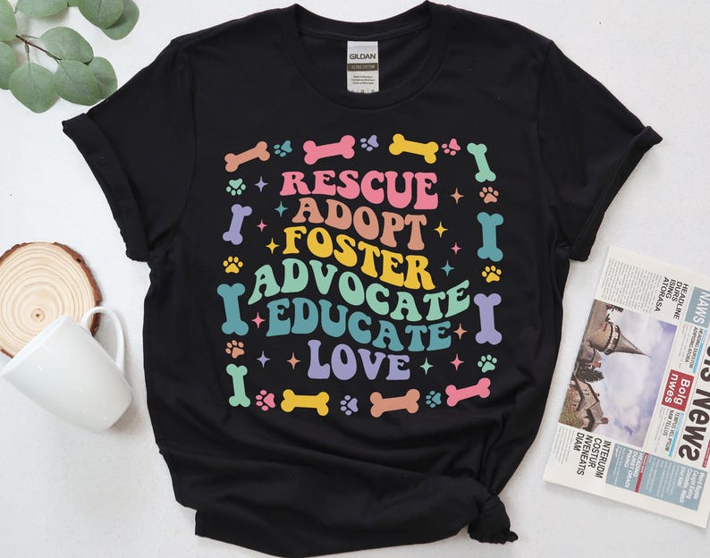May include: Black t-shirt featuring the words "RESCUE, ADOPT, FOSTER, ADVOCATE, EDUCATE, LOVE" in a vibrant, retro style. The design incorporates paw prints, bones, and stars. The shirt is made of a soft fabric.