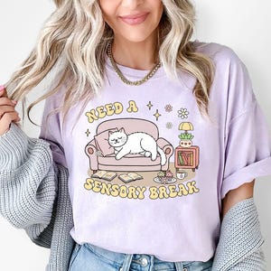 May include: A light purple t-shirt with the text "NEED A SENSORY BREAK" in yellow. The graphic features a white cat lounging on a pink couch, with books, a lamp, and a small table. The shirt is a relaxed fit.