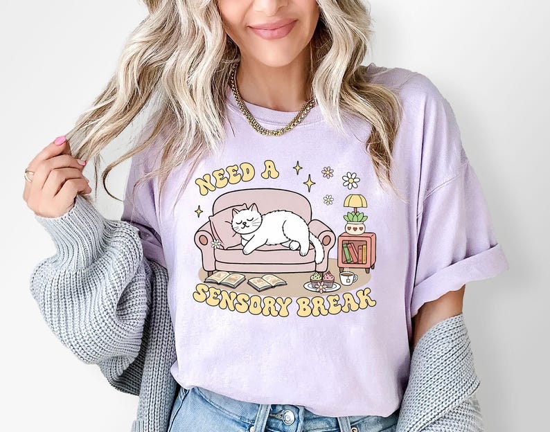 May include: A light purple t-shirt with the text "NEED A SENSORY BREAK" in yellow. The graphic features a white cat lounging on a pink couch, with books, a lamp, and a small table. The shirt is a relaxed fit.