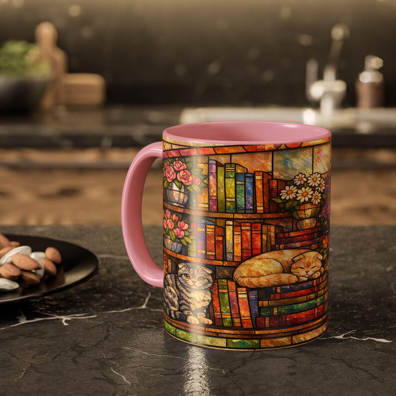Puede incluir: Taza de cer&aacute;mica con asa e interior rosas. La taza presenta un dise&ntilde;o de vidriera de una estanter&iacute;a con libros, flores y dos gatos. Los gatos son un atigrado gris y un gato naranja durmiendo.