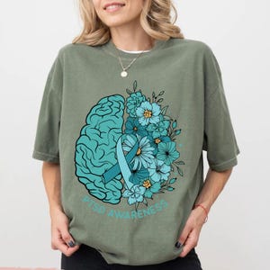May include: Sage green t-shirt with a teal graphic. The graphic features a teal brain, a teal ribbon, and teal flowers. The words "PTSD AWARENESS" are printed in teal below the graphic.