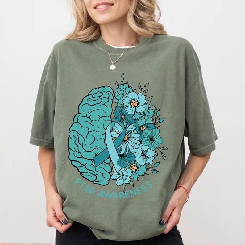 May include: Sage green t-shirt with a teal graphic. The graphic features a teal brain, a teal ribbon, and teal flowers. The words "PTSD AWARENESS" are printed in teal below the graphic.
