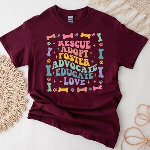 May include: A maroon t-shirt featuring the words "RESCUE ADOPT FOSTER ADVOCATE EDUCATE LOVE" in a colourful, retro font. The design includes paw prints and bone shapes. This is a casual wear item.