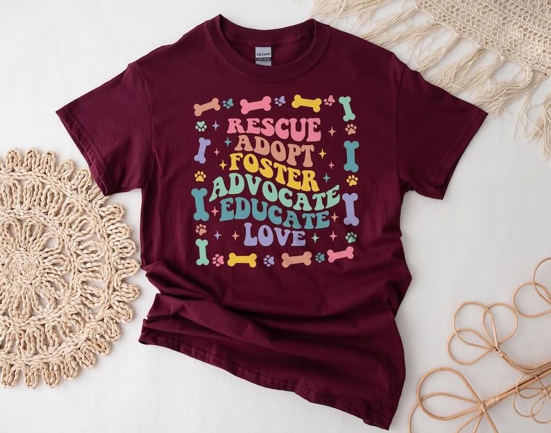 May include: A maroon t-shirt featuring the words "RESCUE ADOPT FOSTER ADVOCATE EDUCATE LOVE" in a colourful, retro font. The design includes paw prints and bone shapes. This is a casual wear item.