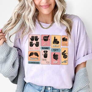 May include: A light purple t-shirt featuring a grid of sign language illustrations. Each square displays a hand gesture with a corresponding word, including "Help," "Please," and "Thank You." The shirt is a soft, casual style.