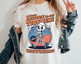 May include: Cream-colored t-shirt featuring a cartoon skeleton with the text "I have Issues in my TISSUES!" and "Ehlers-Danlos Syndrome HAUNTED BY HYPERMOBILITY". The skeleton is inside a blue and orange circle with a checkered border.