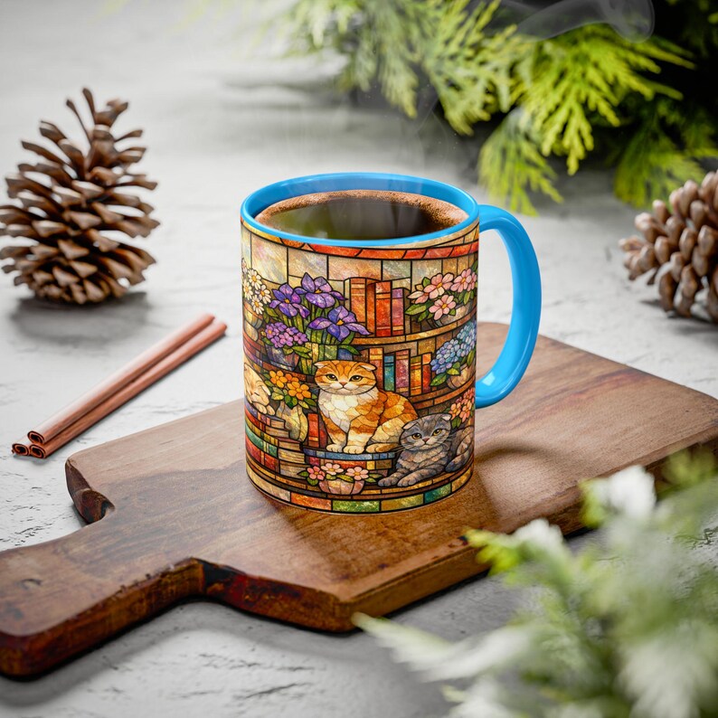 Puede incluir: Taza de caf&eacute; de cer&aacute;mica con un dise&ntilde;o de vidriera que presenta dos gatos, flores y libros. La taza tiene un asa y un borde azules y est&aacute; llena de caf&eacute; caliente. La taza est&aacute; sobre una tabla de cortar de madera.