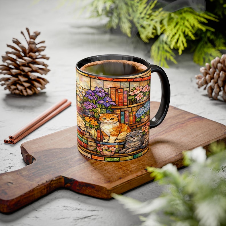 May include: A ceramic coffee mug with a black handle and rim, featuring a stained-glass design of a bookshelf, flowers, and two cats. The mug is filled with coffee and sits on a wooden cutting board.