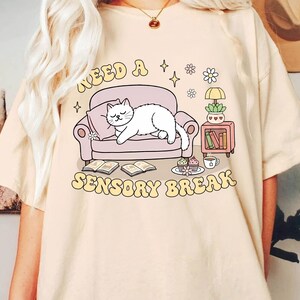 May include: Beige t-shirt with a graphic of a white cat sleeping on a pink couch. The text "NEED A SENSORY BREAK" is in a retro font. The design includes books, a side table, and a cup of coffee.