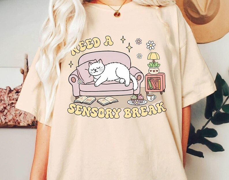 May include: Beige t-shirt with a graphic of a white cat sleeping on a pink couch. The text "NEED A SENSORY BREAK" is in a retro font. The design includes books, a side table, and a cup of coffee.