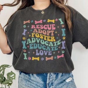 May include: Dark grey t-shirt featuring the words "Rescue, Adopt, Foster, Advocate, Educate, Love" in a rainbow-colored design. The graphic includes paw prints and bone shapes. The shirt has a relaxed, oversized fit.