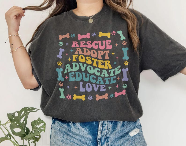 May include: Dark grey t-shirt featuring the words "Rescue, Adopt, Foster, Advocate, Educate, Love" in a rainbow-colored design. The graphic includes paw prints and bone shapes. The shirt has a relaxed, oversized fit.