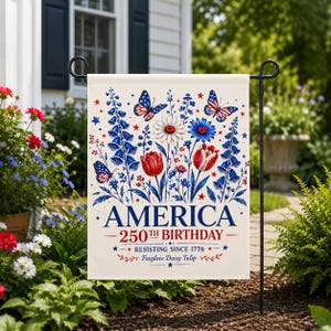 May include: A garden flag with a floral design in red, white, and blue, featuring butterflies and flowers. The flag reads "AMERICA 250TH BIRTHDAY - RESISTING SINCE 1776". The flag is displayed in a garden setting.