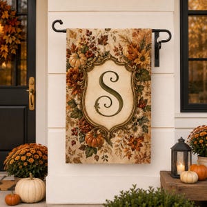 May include: A rectangular garden flag hanging from a black metal stand, featuring a central ornate gold frame enclosing a large dark green letter S. The background is decorated with autumn foliage, orange pumpkins, and red berries in warm tones.