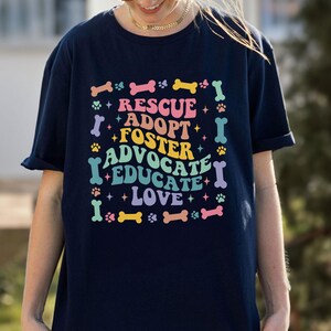 May include: Dark blue t-shirt featuring the words "RESCUE, ADOPT, FOSTER, ADVOCATE, EDUCATE, LOVE" in a colourful design with dog bones, paw prints, and stars. The shirt is a casual, short-sleeved style, perfect for dog lovers.