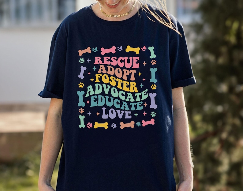 May include: Dark blue t-shirt featuring the words "RESCUE, ADOPT, FOSTER, ADVOCATE, EDUCATE, LOVE" in a colourful design with dog bones, paw prints, and stars. The shirt is a casual, short-sleeved style, perfect for dog lovers.