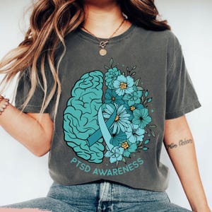 May include: Gray t-shirt featuring a teal brain graphic, a teal ribbon, and teal and blue flowers. The words "PTSD Awareness" are printed in teal below the graphic. The shirt is a comfortable, casual style.