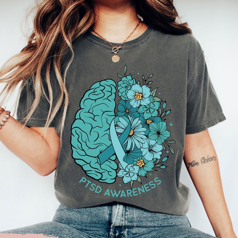 May include: Gray t-shirt featuring a teal brain graphic, a teal ribbon, and teal and blue flowers. The words "PTSD Awareness" are printed in teal below the graphic. The shirt is a comfortable, casual style.