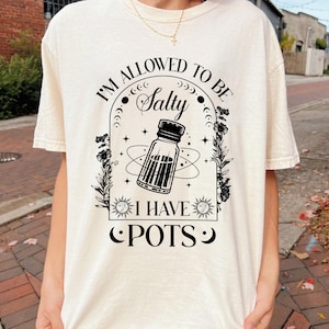 May include: A cream-coloured t-shirt with a black graphic. The design includes a bottle and the text "I'M ALLOWED TO BE Salty I HAVE POTS" in an arch. The shirt has short sleeves and a crew neck.