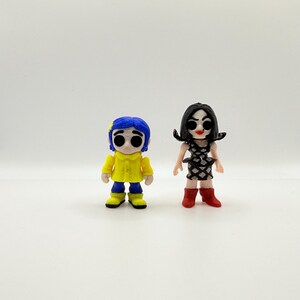 K&ouml;nnte beinhalten: Zwei kleine Figuren von Charakteren aus dem Animationsfilm "Coraline". Eine Figur tr&auml;gt einen gelben Mantel und blaue Haare, die andere ein schwarz-wei&szlig;es Kleid und rote Stiefel. Beide Figuren haben gro&szlig;e, ausdrucksstarke Augen.
