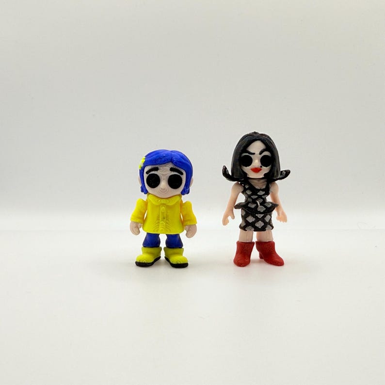 K&ouml;nnte beinhalten: Zwei kleine Figuren von Charakteren aus dem Animationsfilm "Coraline". Eine Figur tr&auml;gt einen gelben Mantel und blaue Haare, die andere ein schwarz-wei&szlig;es Kleid und rote Stiefel. Beide Figuren haben gro&szlig;e, ausdrucksstarke Augen.