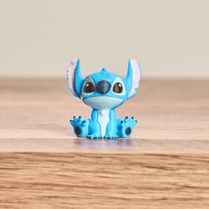 Peut inclure: Petite figurine bleue du personnage Stitch du film Disney Lilo & Stitch. La figurine est assise, les jambes tendues, avec de grandes oreilles roses &agrave; l'int&eacute;rieur. Elle a des yeux noirs et un visage bleu clair.