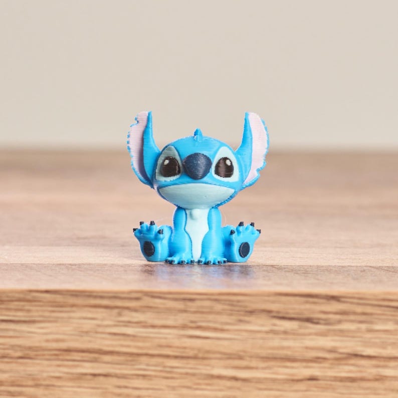 Peut inclure: Petite figurine bleue du personnage Stitch du film Disney Lilo & Stitch. La figurine est assise, les jambes tendues, avec de grandes oreilles roses &agrave; l'int&eacute;rieur. Elle a des yeux noirs et un visage bleu clair.