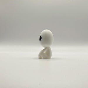 May include: A small, white figurine of a seated humanoid figure with a large, oval head and a single, black eye. The figure is in a seated position, with its knees drawn up. The overall design is minimalist and smooth, with a matte finish.