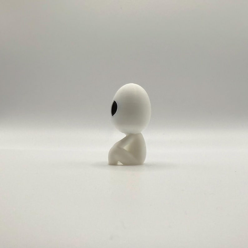 May include: A small, white figurine of a seated humanoid figure with a large, oval head and a single, black eye. The figure is in a seated position, with its knees drawn up. The overall design is minimalist and smooth, with a matte finish.