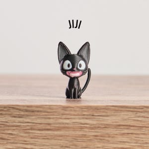May include: A black cat figurine with large, round eyes and a pink mouth. The cat is sitting upright with its tail curved. The word "JIJI" is printed above the cat. The figurine is on a wooden surface.
