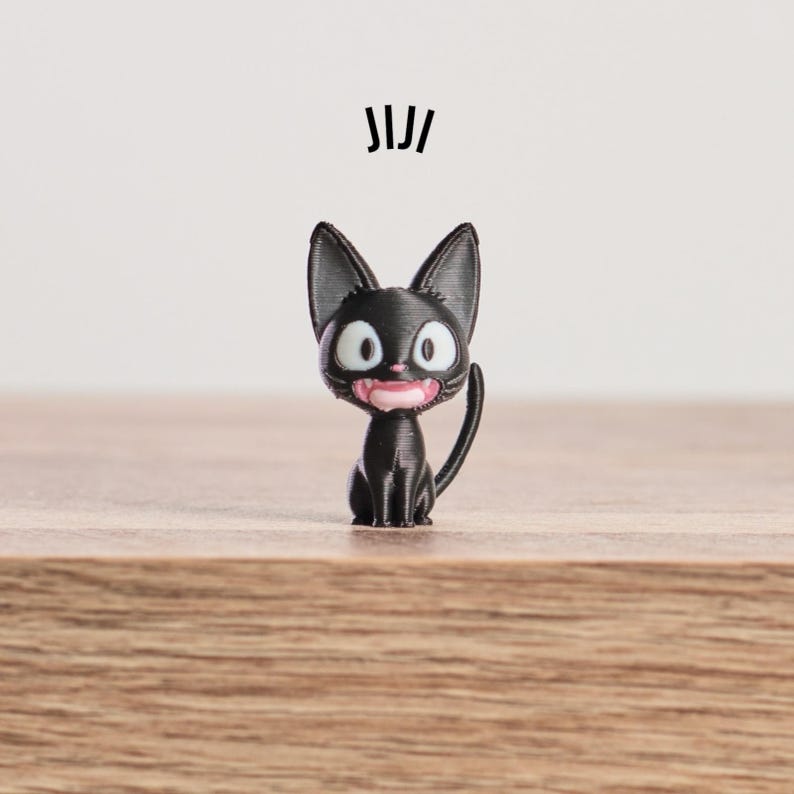 May include: A black cat figurine with large, round eyes and a pink mouth. The cat is sitting upright with its tail curved. The word "JIJI" is printed above the cat. The figurine is on a wooden surface.