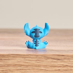 Peut inclure: Petite figurine bleue clair du personnage Stitch du film Disney Lilo & Stitch. La figurine a de grandes oreilles, des marques noires et est assise, les bras et les jambes tendus. Sur une surface en bois.