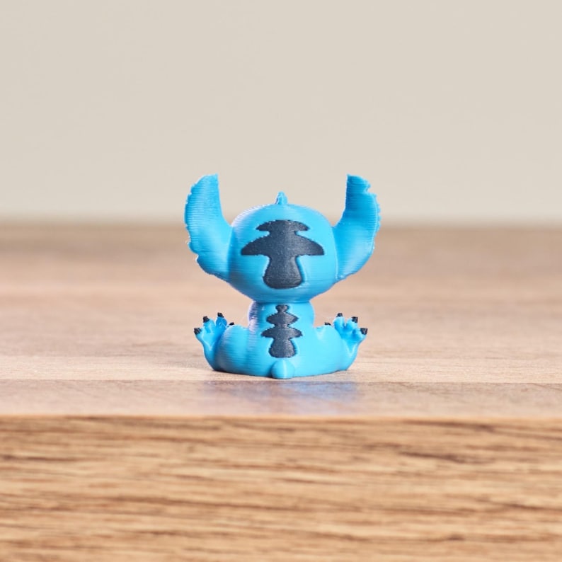 Peut inclure: Petite figurine bleue clair du personnage Stitch du film Disney Lilo & Stitch. La figurine a de grandes oreilles, des marques noires et est assise, les bras et les jambes tendus. Sur une surface en bois.