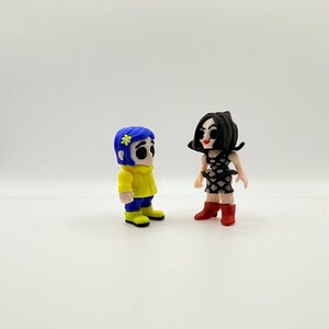 K&ouml;nnte beinhalten: Zwei kleine Figuren von Filmfiguren. Eine Figur hat blaue Haare, eine gelbe Jacke, blaue Hosen und gelbe Stiefel. Die andere Figur hat schwarze Haare, ein schwarz-wei&szlig;es Kleid und rote Stiefel.